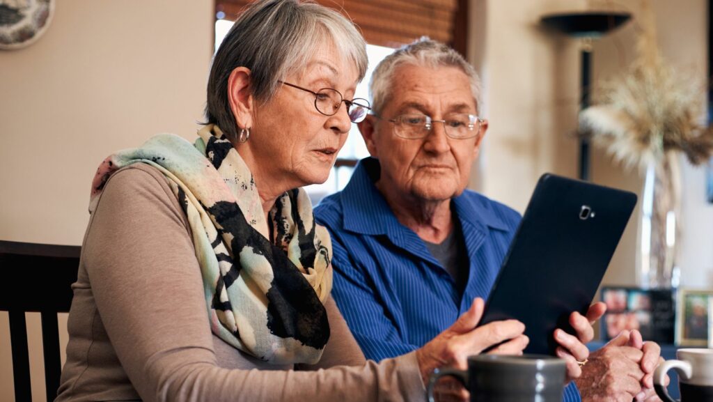 An elderly couple sitting at a table, attentively looking at a tablet device. The woman is wearing glasses and a colorful scarf, while the man in a blue shirt holds a coffee mug. They appear focused and engaged, possibly learning or reviewing something on the screen.