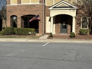 Brick building with an American flag and a colorful hanging decoration on the left unit, and a doorway with a small porch on the right unit, viewed from the parking lot.