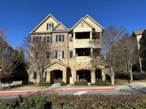 Three-story residential building with stone and brick exterior, featuring balconies, arched entrances, and surrounded by trees and bushes under a clear blue sky.
