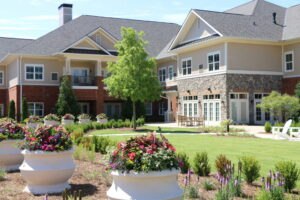Modern assisted living facility with landscaped gardens, large planters filled with flowers, and a mix of brick and stone exterior.