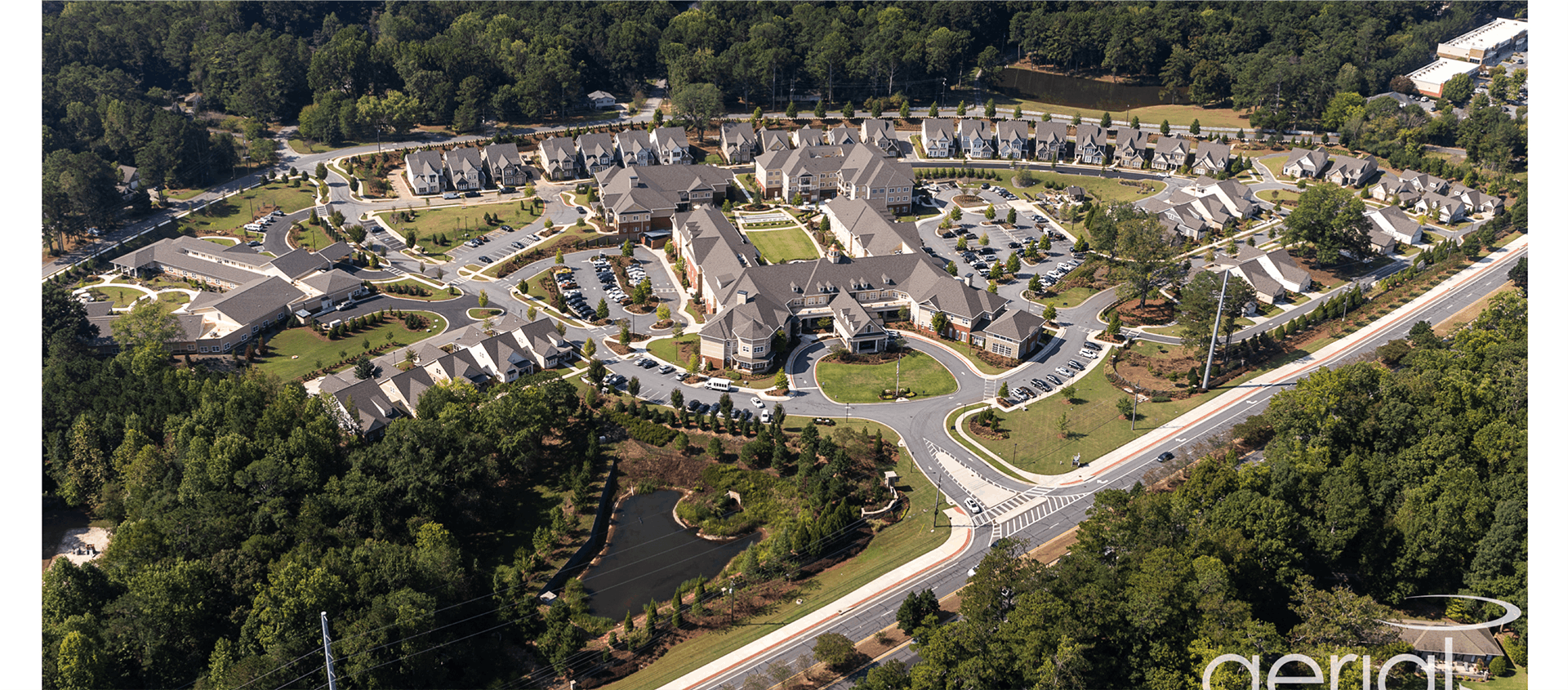 Aerial view of a large residential complex surrounded by trees, featuring multiple buildings, roads, landscaped areas, and a small pond.