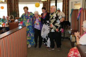 Four people in Hawaiian-themed outfits and leis walk arm-in-arm at an indoor event, while others sit and watch.