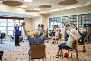 A group of seniors sit in chairs raising dumbbells while following an instructor during an exercise class in a well-lit room.