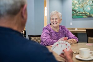 An elderly woman smiles while playing cards with another person at a table in a bright room with art on the wall.