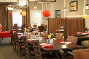 A dining area with brown tables and chairs, striped booth seating, floral centerpieces, red napkins, and hanging paper lantern decorations.