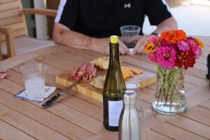 A person sits at a wooden table with a bottle of wine, a water bottle, a platter of meat and cheese, a glass of water, and a vase of colorful flowers.