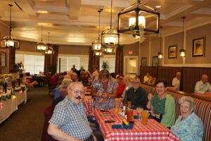 A group of elderly people sit at tables with red checkered tablecloths in a well-lit dining room, some smiling at the camera.