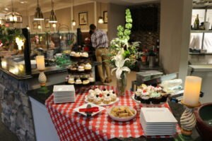 A dessert buffet with cupcakes, tarts, and cookies on a red checkered tablecloth, surrounded by plates, candles, and a flower arrangement in a restaurant setting.