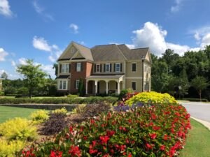 A two-story suburban house with brick and beige siding, surrounded by a green lawn and colorful flower beds under a blue sky with scattered clouds.