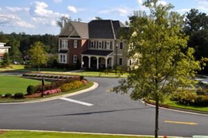 A large two-story suburban house with a manicured lawn, colorful flowerbeds, and a circular driveway on a sunny day.