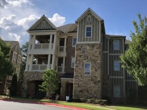 Three-story apartment building with brick and stone facade, covered balconies, and tall windows, surrounded by greenery under a partly cloudy sky.