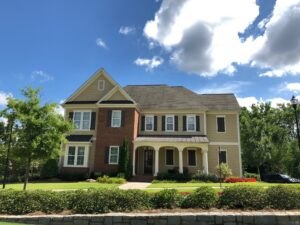 Two-story suburban house with brick and beige siding, shuttered windows, front porch, and landscaped yard under a partly cloudy sky.