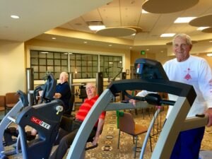 Three elderly men exercise on cardio machines in a well-lit gym with beige walls and ceiling lights; two are seated, one is standing and smiling at the camera.