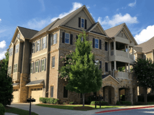 Three-story townhouse with brick and siding exterior, balconies, multiple windows, and attached garage, surrounded by trees and landscaping under a blue sky.