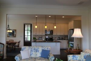 Open-concept living room and kitchen with pendant lights, cream cabinets, stainless steel appliances, and floral-patterned sofas; dining area visible in the background.