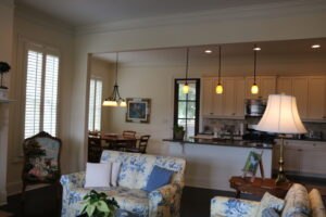 A bright, open living room with floral-patterned sofas, a wooden coffee table, and a view into a kitchen with hanging pendant lights and dining area.
