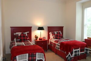 A bedroom with two twin beds featuring red headboards and plaid bedding, a red nightstand with a lamp and clock between the beds, and a window with blinds on the right.