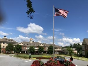 An American flag on a tall flagpole waves in front of a large brick building with a manicured lawn and flower beds under a blue sky with scattered clouds.