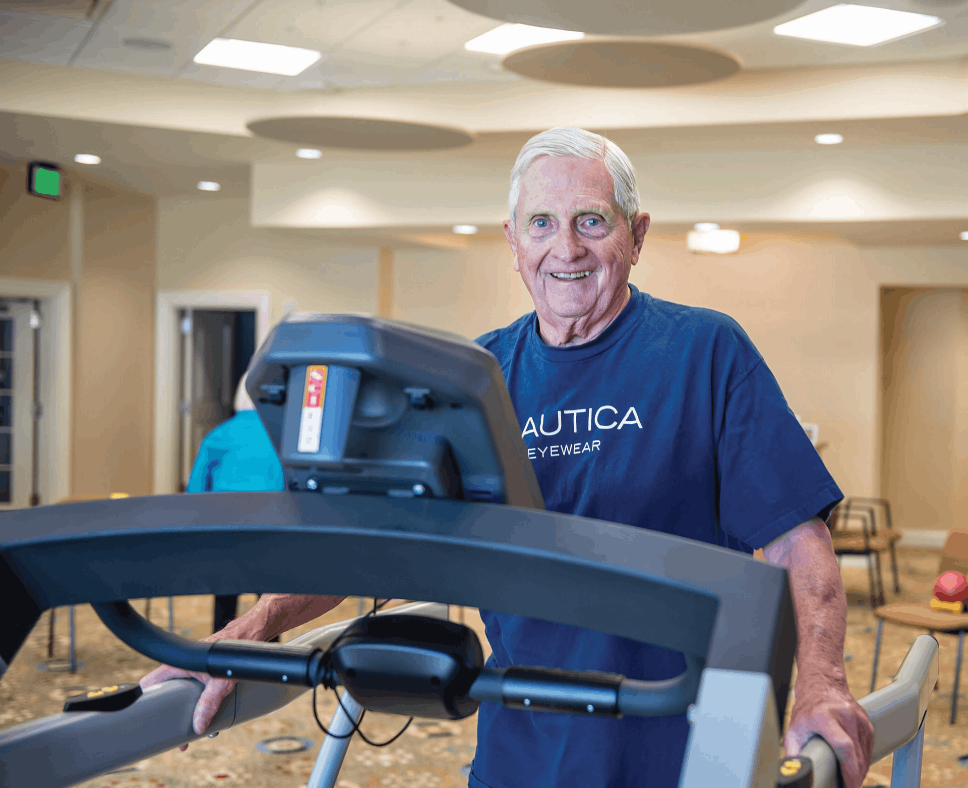 An older man in a blue T-shirt stands on a treadmill in a well-lit indoor space, smiling at the camera.