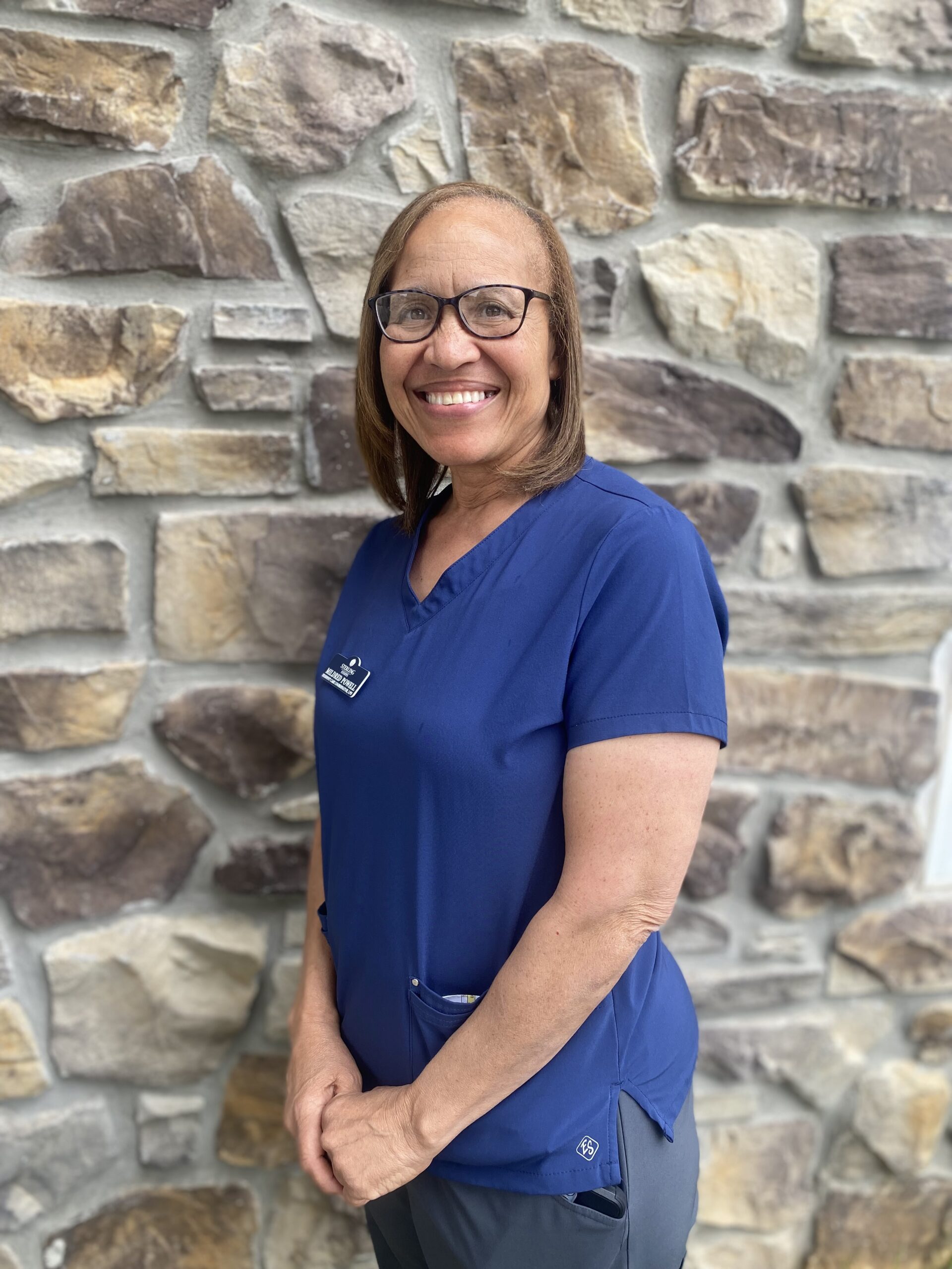 A woman wearing glasses and blue scrubs stands and smiles in front of a stone wall.