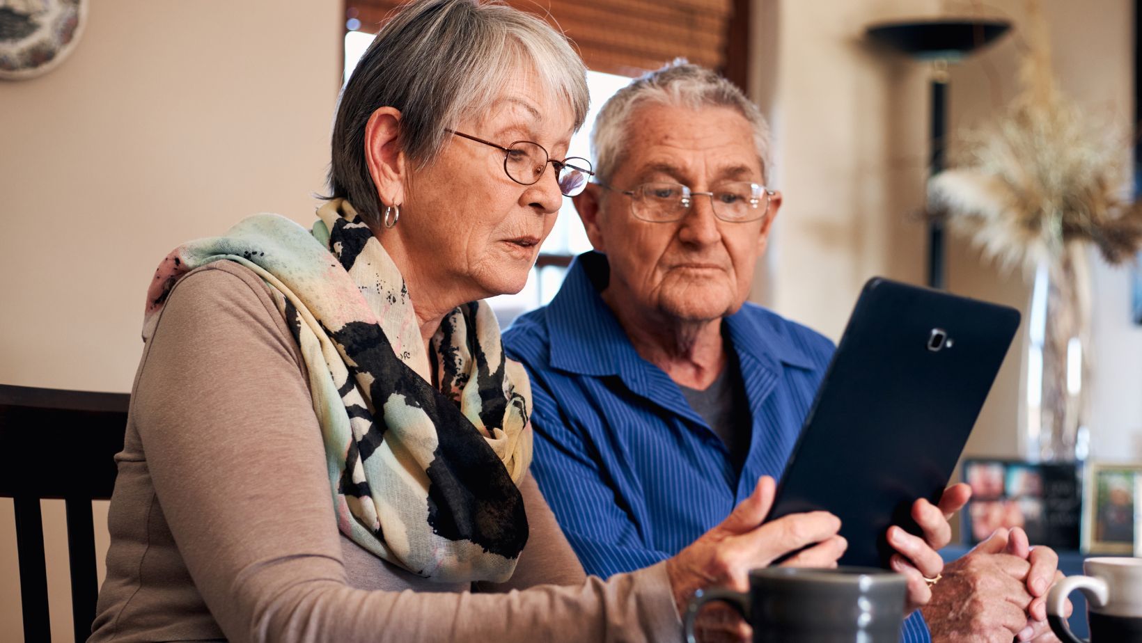 An elderly couple sitting at a table, attentively looking at a tablet device. The woman is wearing glasses and a colorful scarf, while the man in a blue shirt holds a coffee mug. They appear focused and engaged, possibly learning or reviewing something on the screen.