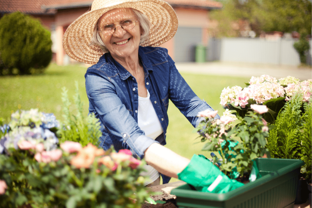 An older woman enjoying Independent Living wears a straw hat and green gloves while gardening outdoors, smiling as she tends to flowers in a planter box.