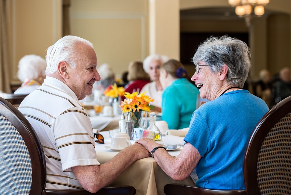 Two older adults sit at a dining table holding hands and smiling at each other, with other people and flowers visible in the background.