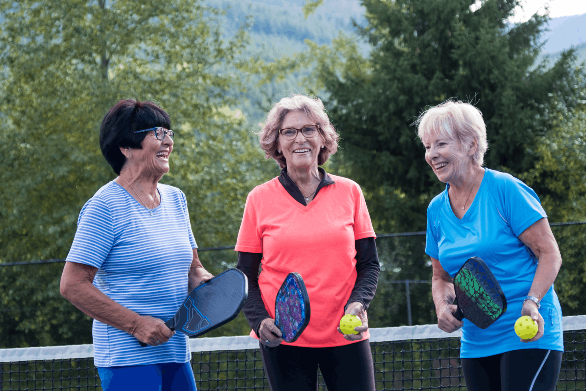 Three older women stand outdoors on a pickleball court, each holding a paddle and ball, smiling in athletic clothing. The vibrant scene reflects the active lifestyle and camaraderie found in Independent Living communities. Trees and a net appear in the background.