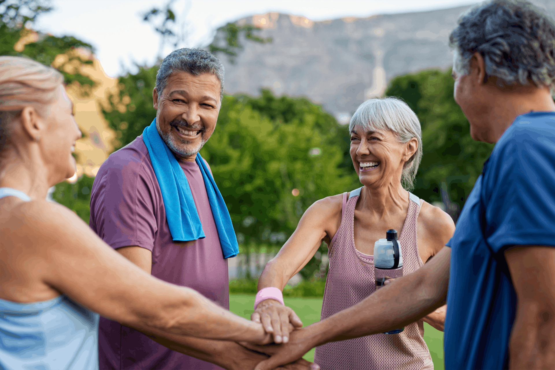 Four older adults stand outdoors in athletic wear, smiling and stacking their hands together in a gesture of teamwork—a perfect example of the camaraderie found in Independent Living. Trees and mountains are visible in the background.