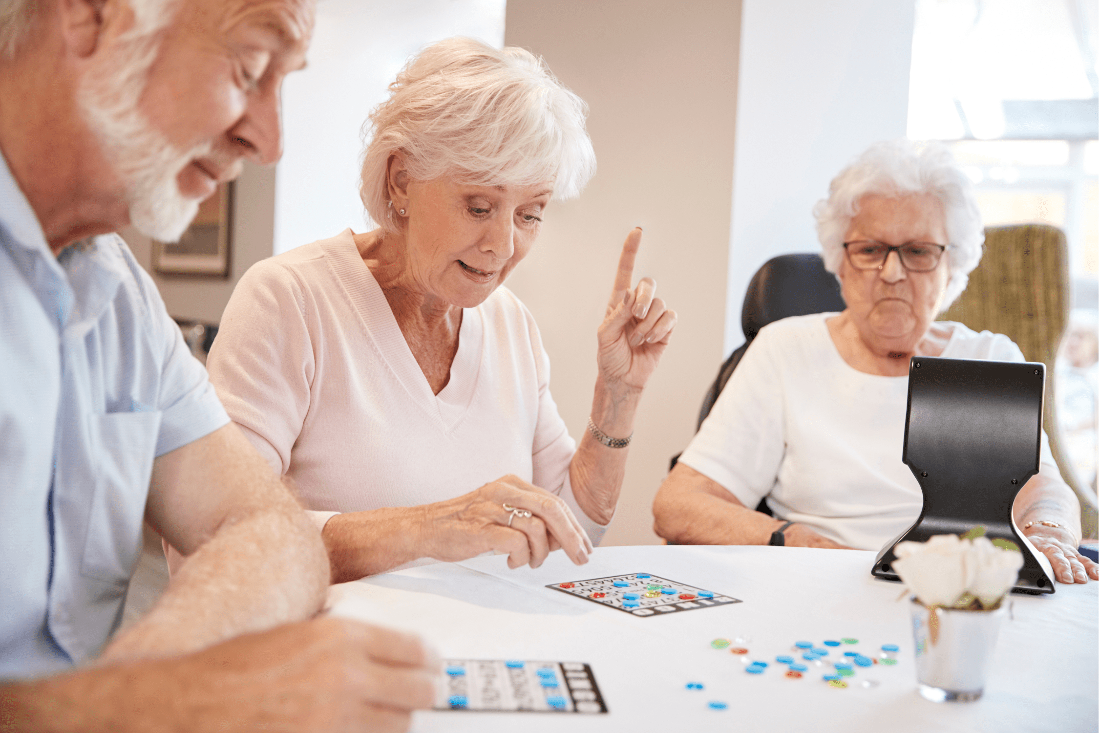 Three older adults enjoy a lively game with cards and colored chips in an Independent Living community; one woman raises her finger while the others focus on the fun.